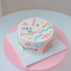 a white cake with rainbows and clouds on it sitting on a pink plate next to a book