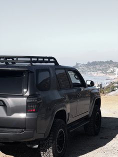 a gray truck parked on top of a dirt road next to the ocean and mountains