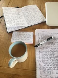 a cup of coffee next to an open notebook and pen on a wooden table top