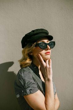 a woman wearing sunglasses and a hat with her hands on her face, leaning against a wall