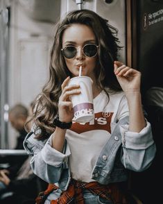 a woman wearing sunglasses and drinking from a paper cup while sitting on a subway train