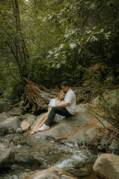 a man and woman sitting next to each other on rocks near a river in the woods