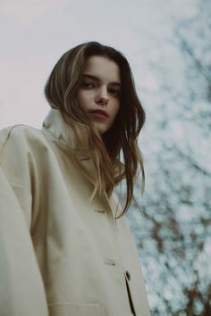 a woman with long hair standing in front of trees wearing a trench coat and looking off into the distance