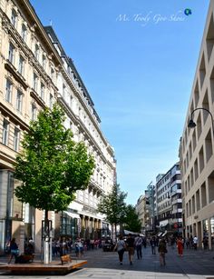 people are walking down the street in front of tall buildings and trees on both sides