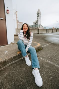 a woman sitting on the curb with her legs crossed and smiling at the camera while wearing white sneakers