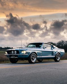 an old mustang car is parked on the side of the road in front of some clouds