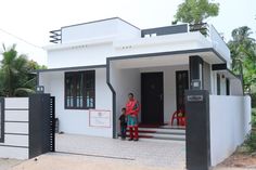 two people standing in front of a small white and black house with red chairs on the porch