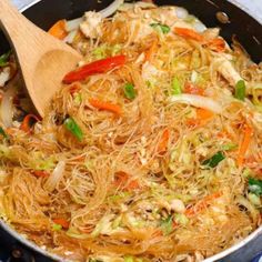 a wok filled with noodles and vegetables on top of a blue table cloth next to a wooden spoon