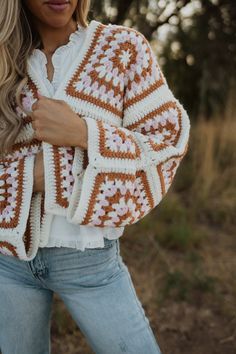 a woman wearing a crochet jacket and jeans standing in the woods with her arms crossed