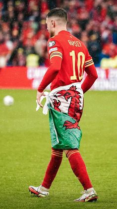 a man in red jersey and green shorts walking on field with soccer ball behind him