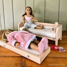 two dolls sitting on top of a doll bed next to a wooden table with a pink blanket