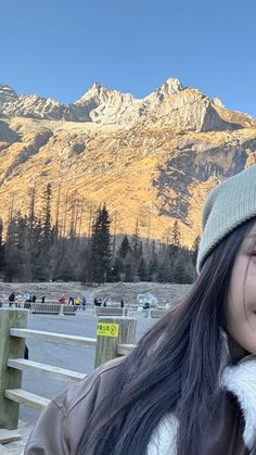 a woman with long hair wearing a beanie and smiling at the camera in front of mountains