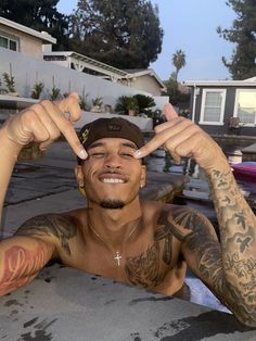 a man with tattoos on his arms and chest sitting in front of a house, making the peace sign