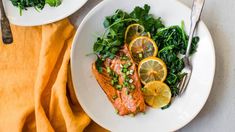 two plates with salmon, spinach and lemons on them next to silverware