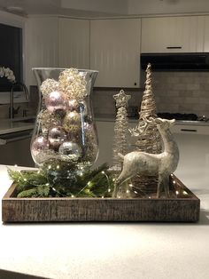 a wooden tray topped with christmas ornaments on top of a kitchen counter next to a glass vase