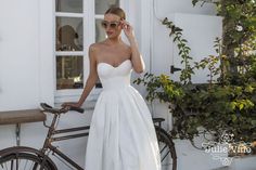 a woman in a white dress standing next to a bike and holding her hand on her head