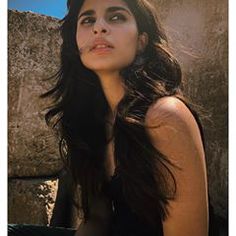 a woman with long black hair sitting next to a stone wall and looking off into the distance