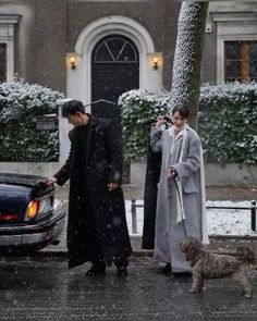 two people and a dog standing in front of a car on a street with snow