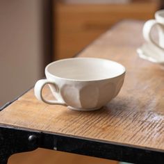 two white cups sitting on top of a wooden table next to each other with saucers