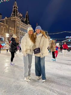 two women standing on an ice rink at night with lights strung above the buildings behind them