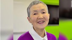 an older woman wearing a purple sweater and white collared shirt smiles at the camera