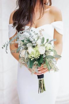 a woman in a white dress holding a bridal bouquet with greenery and flowers