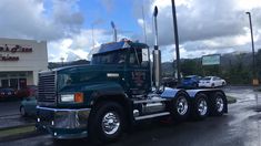 a green semi truck parked in front of a store on a cloudy day with cars behind it