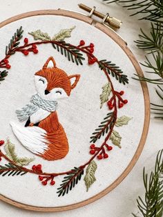 an embroidered hoop with a fox and pine needles on it, surrounded by greenery