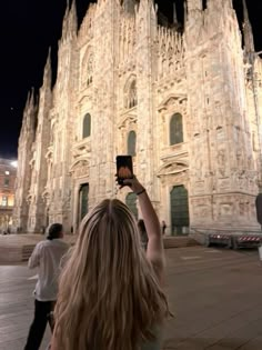 a woman holding up her cell phone in front of a cathedral at night with people walking by