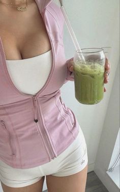 a woman in white shorts and pink jacket holding a glass with green smoothie inside