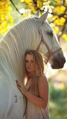 a woman standing next to a white horse under a tree with yellow leaves on it