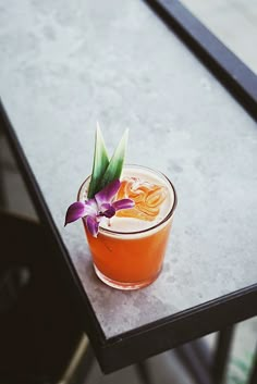 an orange drink with a purple flower on the rim sitting on top of a table