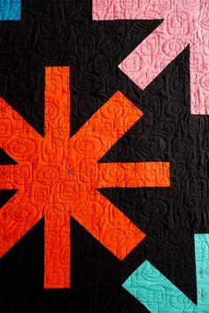 a black quilt with orange and pink arrows on it's center, in the middle of an abstract design