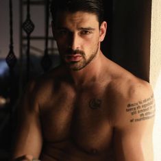 a shirtless man with a tattoo on his left arm and chest, leaning against a wall