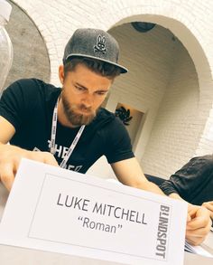 a man sitting at a table with a sign in front of him that says luke mitchell roman