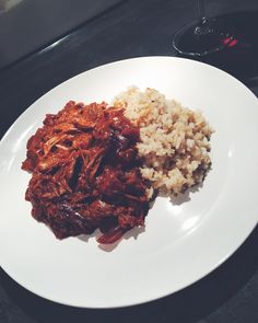 a white plate topped with meat and rice next to a glass of wine on a table