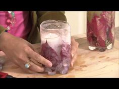 a woman holding a glass filled with ice and some purple flowers in it's hands