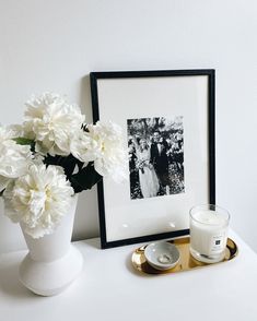 a white vase filled with flowers sitting next to a candle and framed photograph on a table