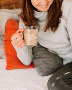 a woman holding a coffee mug while sitting on a bed in front of a pillow