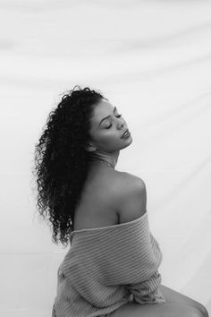 a black and white photo of a woman with curly hair sitting on a stool looking off to the side