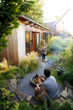 two people sitting around a fire pit in the middle of a yard with trees and plants