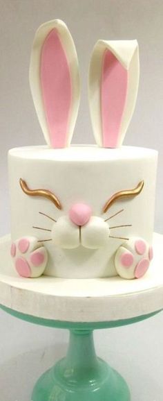 a white cake with pink and gold bunny ears on it's face, sitting on top of a green pedestal