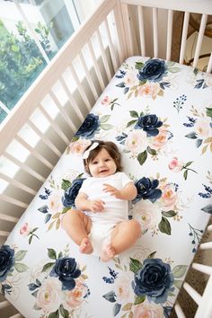 a baby sitting in a crib with flowers on the sheets and smiling at the camera