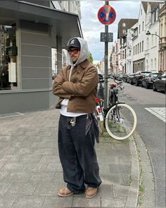 A man in a casual streetwear outfit stands with arms crossed. He's wearing a brown jacket over a gray hoodie, black Yankees cap, loose black pants with a bandana, and brown shoes. The setting is an urban street corner with parked cars and a bike nearby. Looks Hip Hop, Streetwear Inspo, Winter Streetwear, Streetwear Fits, Skater Jeans, Street Style Outfits Men, Street Fashion Men Streetwear, Guys Clothing Styles, Mens Outfit Inspiration