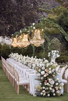 a long table set up with white flowers and chandeliers for an outdoor wedding