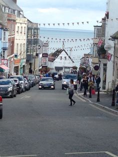 people are walking down the street in front of parked cars and buildings with flags hanging above them