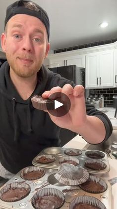 a man holding up a chocolate cupcake in front of his muffin tins