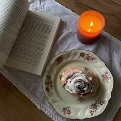 an open book on a table next to a plate with a pastry and a candle