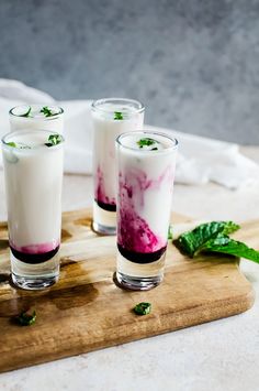 three glasses filled with liquid sitting on top of a wooden cutting board next to mint leaves