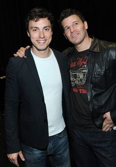 two men standing next to each other in front of a black curtain and one is wearing a leather jacket